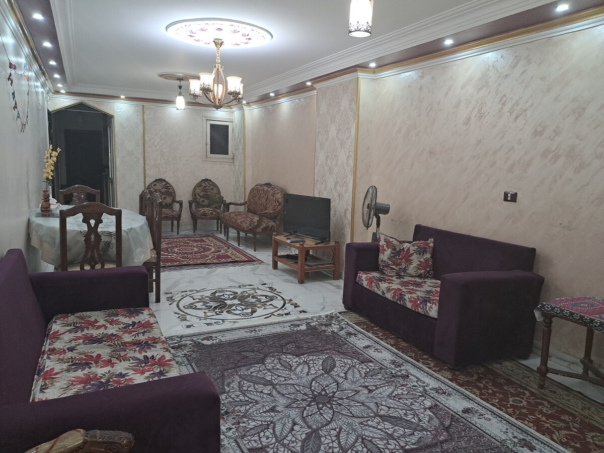 A spacious living area features a patterned area rug centered on a marble floor. Two plush sofas with floral upholstery are accompanied by wooden chairs. A dining table with a decorative cloth is visible, and a television stands against the wall, providing a cozy gathering space.