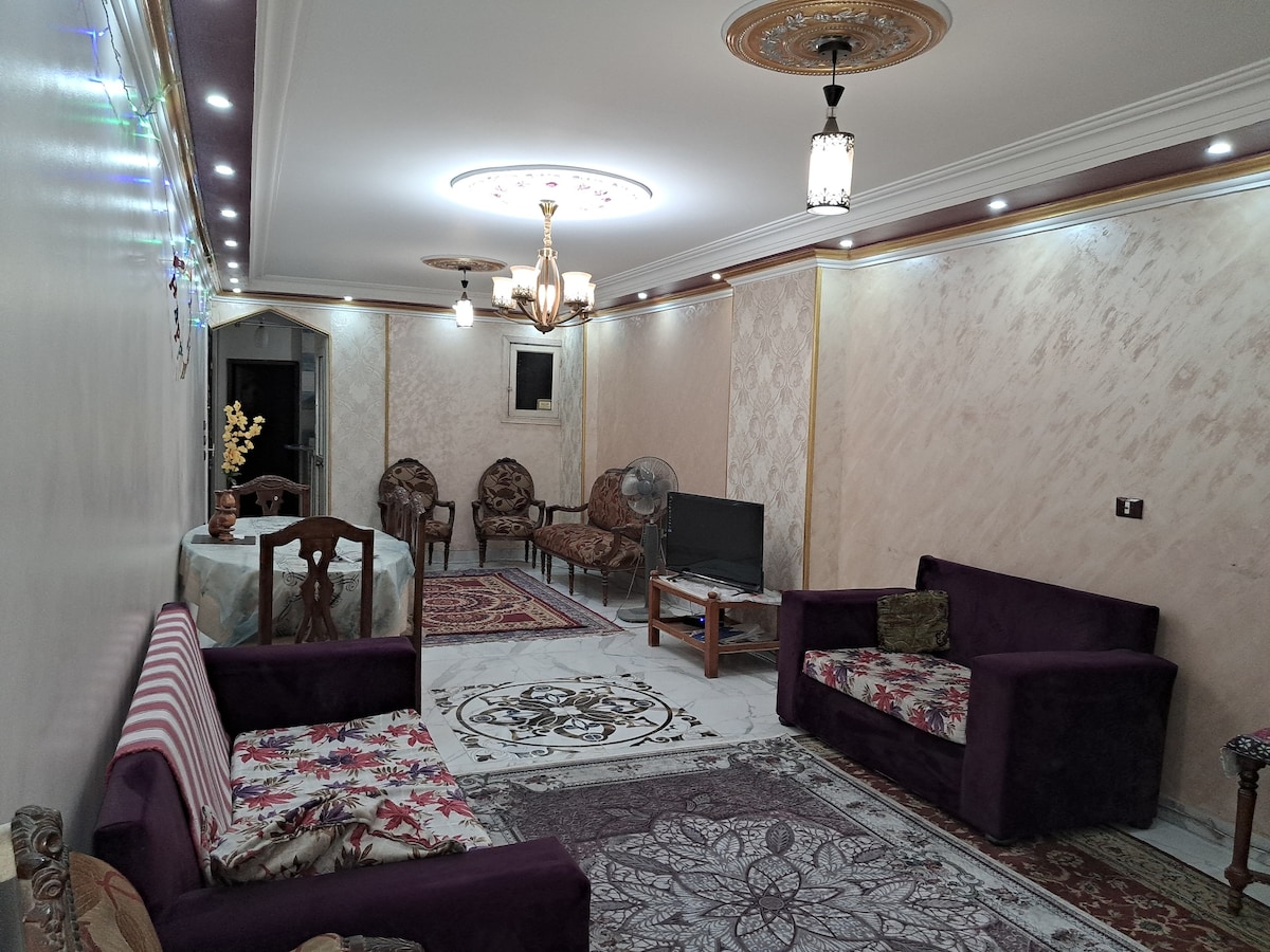 A spacious living area is featured, furnished with plush seating arrangements in rich colors. Decorative elements include a patterned area rug and various light fixtures enhancing the space. A television is positioned against one wall, while a small dining table is nearby.