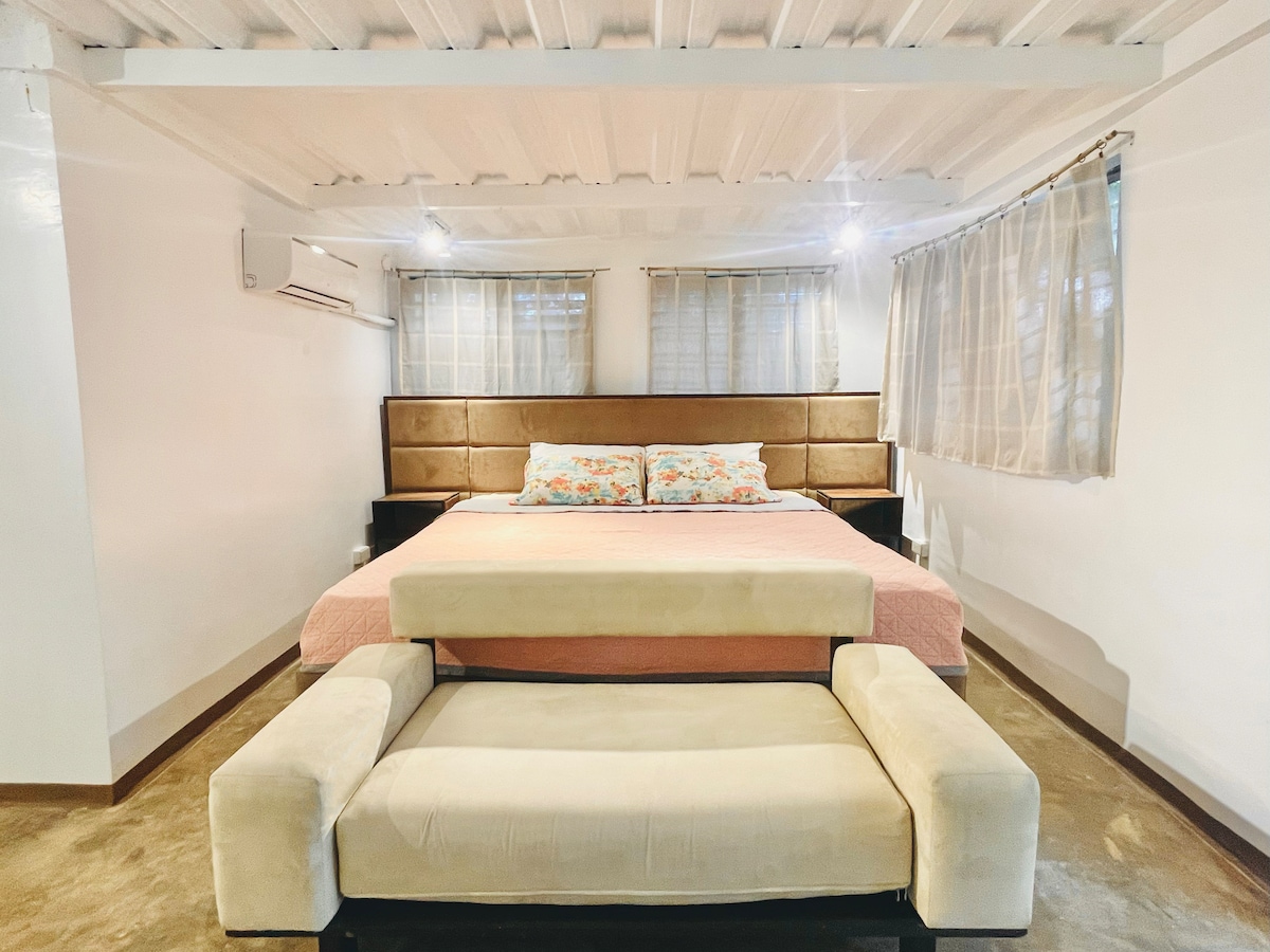 A spacious sleeping area is highlighted by a custom bed larger than king size, featuring soft floral bedding. Two windows offer natural light, while a compact sofa is positioned at the foot of the bed, enhancing the room's functionality.