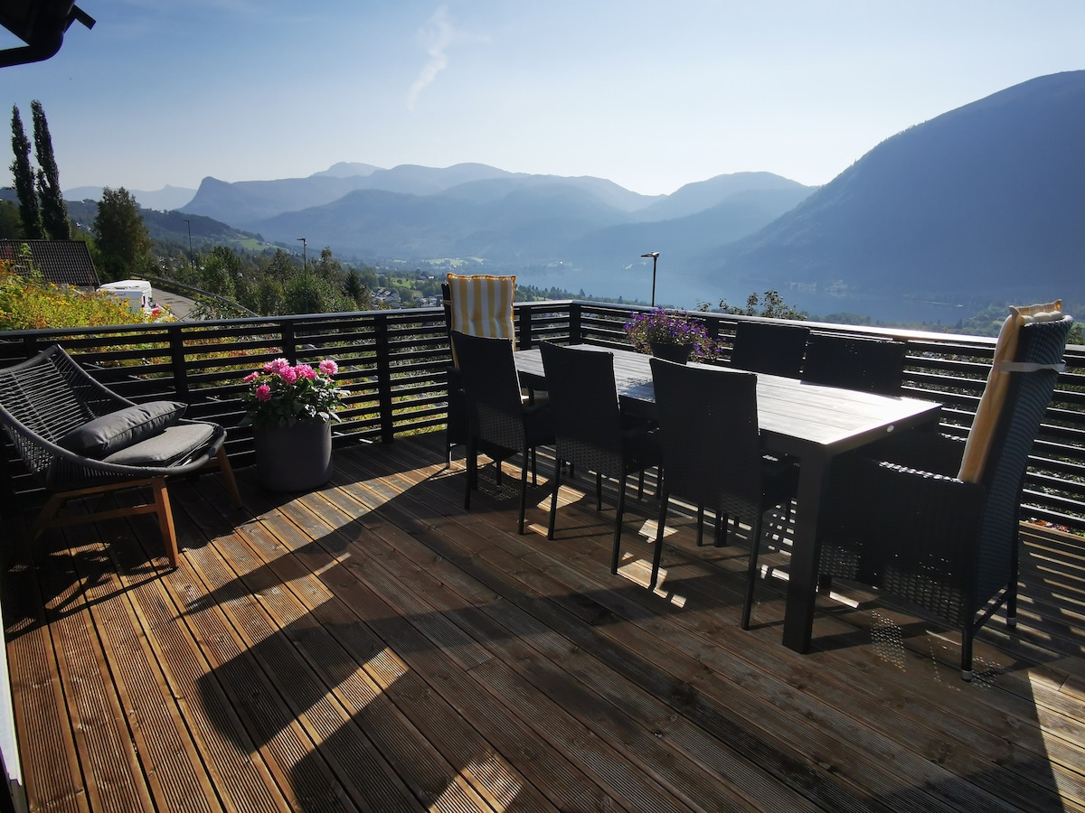 A spacious outdoor terrace features a large dining table surrounded by several chairs, complemented by potted flowers. A lounge chair is positioned nearby. The backdrop consists of majestic mountains under a clear sky, creating a serene setting.