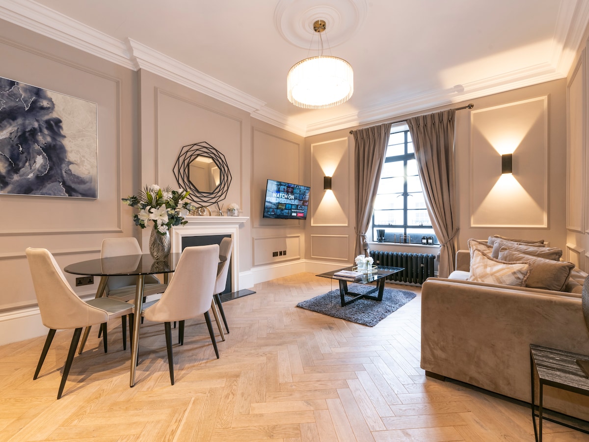 An elegant living area features a plush sofa adorned with decorative cushions and a mounted flat-screen TV. A circular dining table with stylish chairs complements the herringbone-patterned wood floor. Large windows allow natural light to fill the space, enhanced by softly draped curtains.