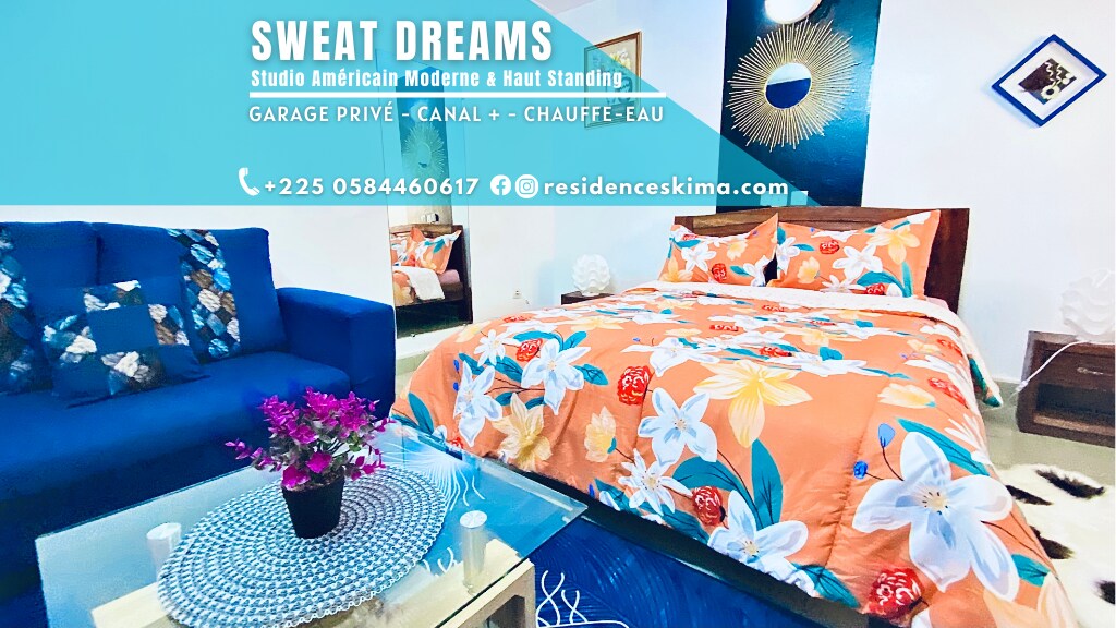 A spacious sleeping area features a brightly colored bedspread adorned with floral patterns. A cozy blue sofa is positioned nearby, complemented by a small vase of purple flowers. Soft lighting enhances the room's inviting ambiance, while decorative elements add character.