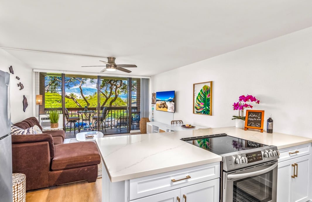 A spacious kitchen features modern white cabinetry and a large island with quartz countertops. Stainless steel appliances are visible, including an oven and refrigerator. The living area, partially seen in the background, includes a cozy couch and access to a balcony with views of lush greenery.