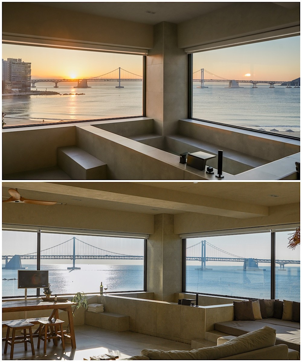 Large windows frame views of the Gwangan Bridge and the sea, showcasing the vibrant colors of sunrise. A relaxing seating area is set by the window, complemented by a dining table nearby, both designed to enhance the experience of the beautiful landscape.