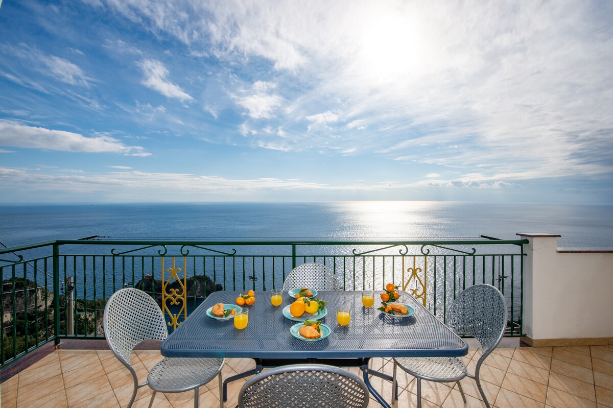 A spacious terrace features a dining table set for four, adorned with fruit and breakfast items. The view overlooks the expansive sea, shimmering under bright sunlight. Decorative railing complements the scenic backdrop, while the sky displays a mix of clouds and blue hues.