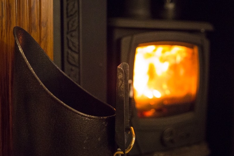 A warm multifuel fire is observed within a dark fireplace, glowing softly with orange and yellow flames. A metal fire tool is positioned to the left, adding a rustic element to the inviting ambiance of the space.