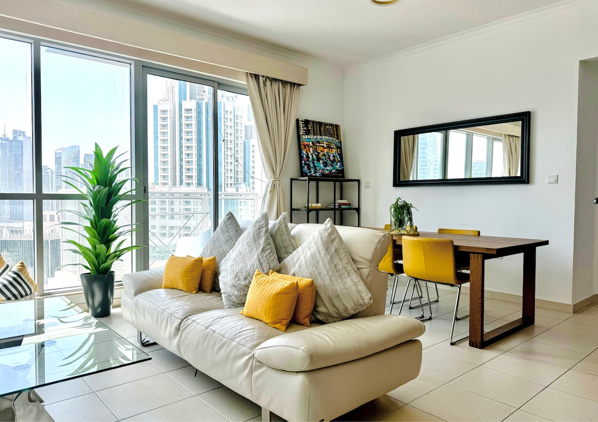 A spacious living area is highlighted by large windows, allowing natural light to fill the space. A comfortable white sofa is adorned with plush pillows, accompanied by a glass coffee table. A dining table with four yellow chairs complements the modern decor, while greenery adds a touch of warmth.