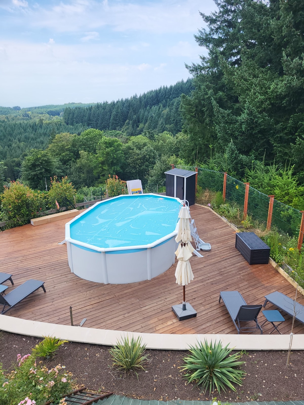 An outdoor area features a circular swimming pool surrounded by a wooden deck. Sun loungers are positioned around the pool, accompanied by a shaded umbrella. Lush greenery and trees provide a natural backdrop, enhancing the tranquil environment.