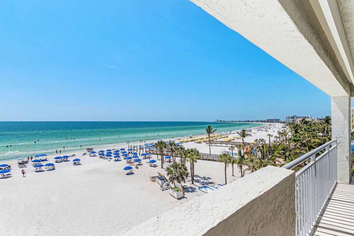 A view from the balcony captures the expansive beach and vibrant turquoise waters. Sun loungers are arranged on the soft, white sand below, framed by gently swaying palm trees. The clear blue sky stretches overhead, enhancing the sense of spaciousness and serenity.