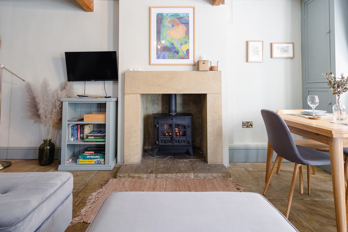 A cozy living area features a gas-burning stove set in a stone fireplace, surrounded by light-colored walls. A small table is positioned nearby, with two chairs and a variety of decorative elements, including books and art, enhancing the space's charm.