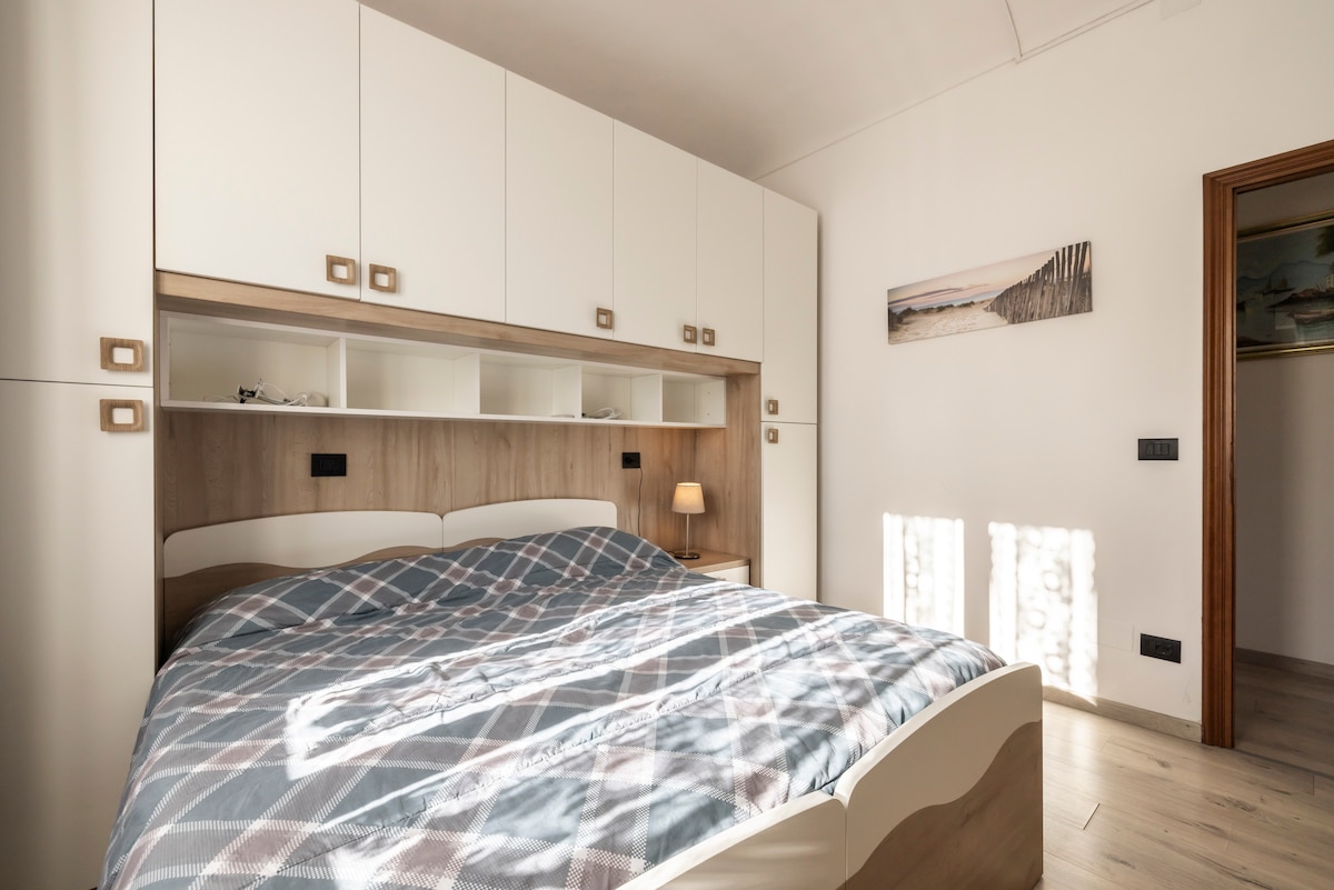 A well-lit bedroom contains a spacious bed with a patterned blanket. Above the bed, ample storage is provided by modern cabinetry. Soft light from a bedside lamp creates a calming atmosphere, while sunlight casts gentle shadows on the floor.