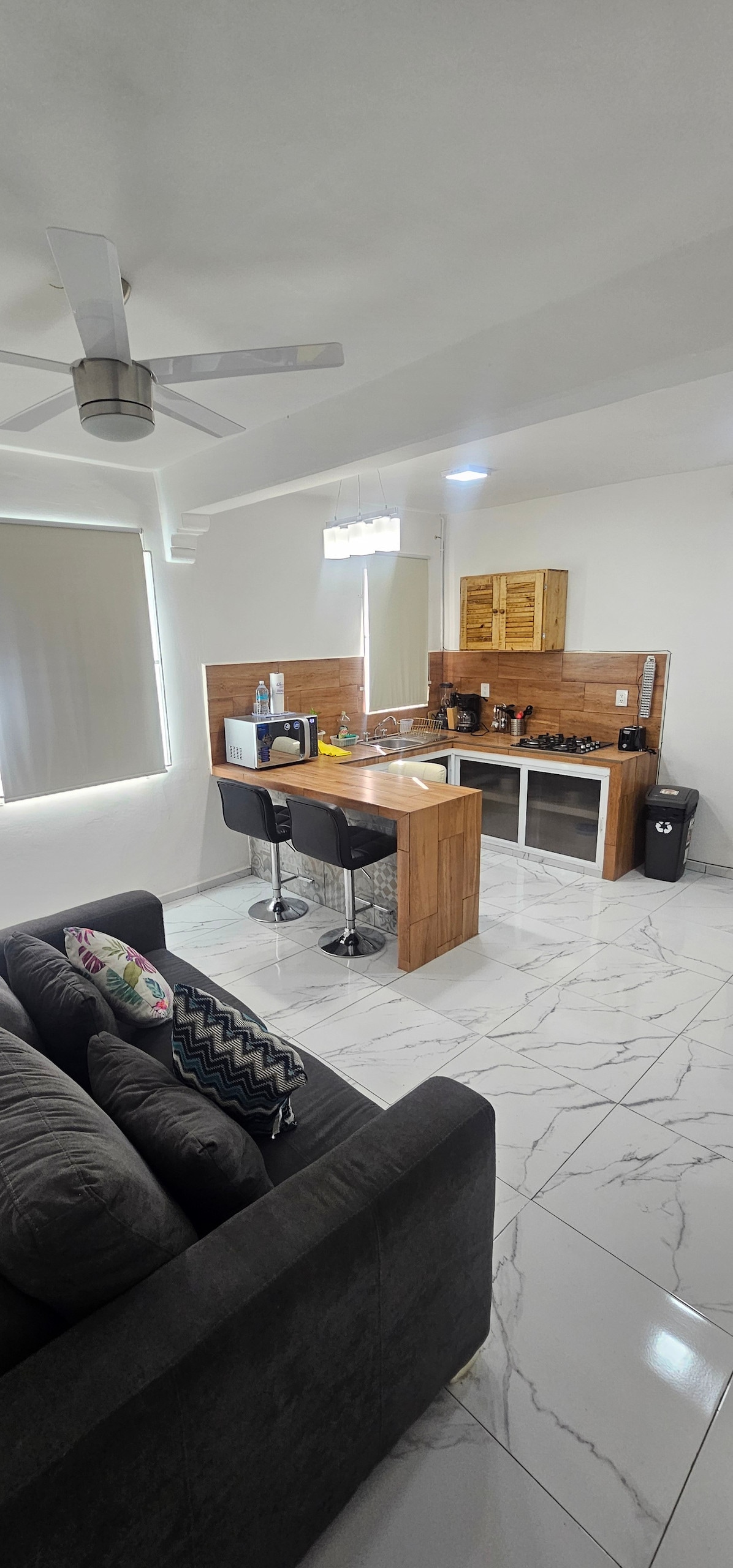 A minimalist kitchen area features wooden cabinetry and modern appliances, including a microwave and coffee maker. Two black bar stools are positioned at a wooden counter, providing space for casual dining. A comfortable gray sofa is visible in the living area, complemented by decorative cushions.