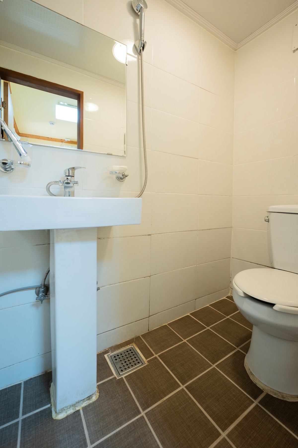 A compact bathroom features white tiled walls and a matching floor. A modern sink with a chrome faucet is fixed above a clean, white pedestal. A toilet is positioned nearby, and a wall-mounted mirror is above the sink, reflecting the simple, functional design.