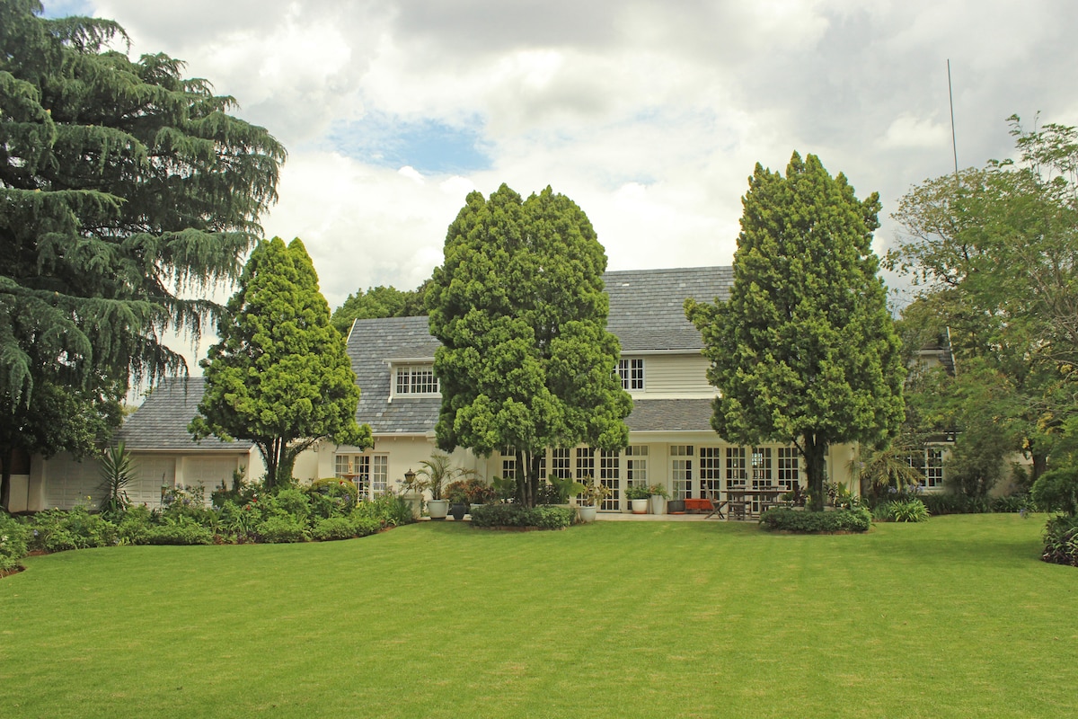 The villa is set amidst manicured lawns and lush greenery, featuring tall trees that frame the exterior. The two-story structure exhibits a classic design with large windows and a slate roof, inviting natural light into the spacious surroundings.