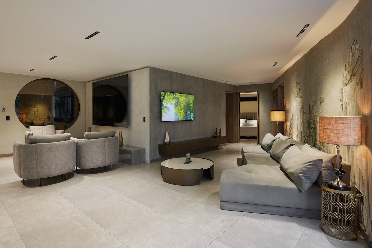 A modern living area features a spacious sectional sofa and two circular armchairs arranged around a circular coffee table. Wall-mounted smart TVs are positioned for viewing, while warm lighting is provided by two decorative lamps. Textured wall designs add visual interest to the neutral decor.