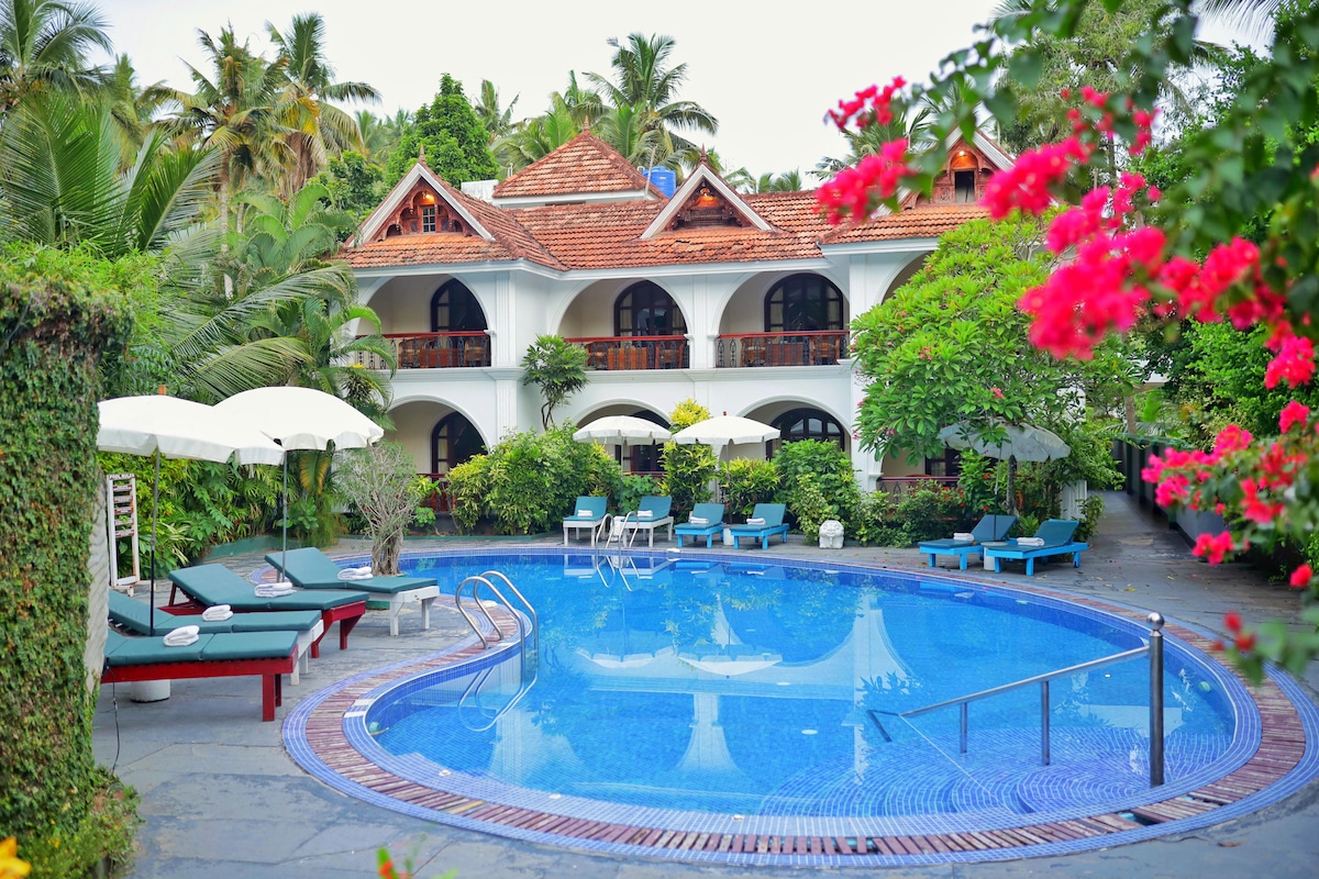 A vibrant pool area is surrounded by lush greenery and colorful flowers. Comfortable sun loungers are positioned around the rounded pool, complemented by white umbrellas. The villa's charming architecture, featuring arched balconies and a red-tiled roof, provides an inviting backdrop.