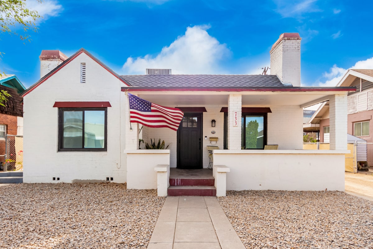 The exterior of a historic bungalow is showcased, featuring a white facade with red accents. An American flag adorns the front, while a walkway leads to the entrance. Gravel surrounds the home, and nearby structures are partially visible against a clear blue sky.