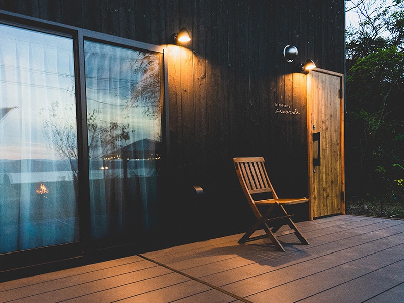 A simple wooden chair is positioned on a deck outside the entrance, framed by large glass doors that reflect the surrounding landscape. Soft lighting highlights the dark wooden facade of the building, creating a serene and inviting atmosphere.