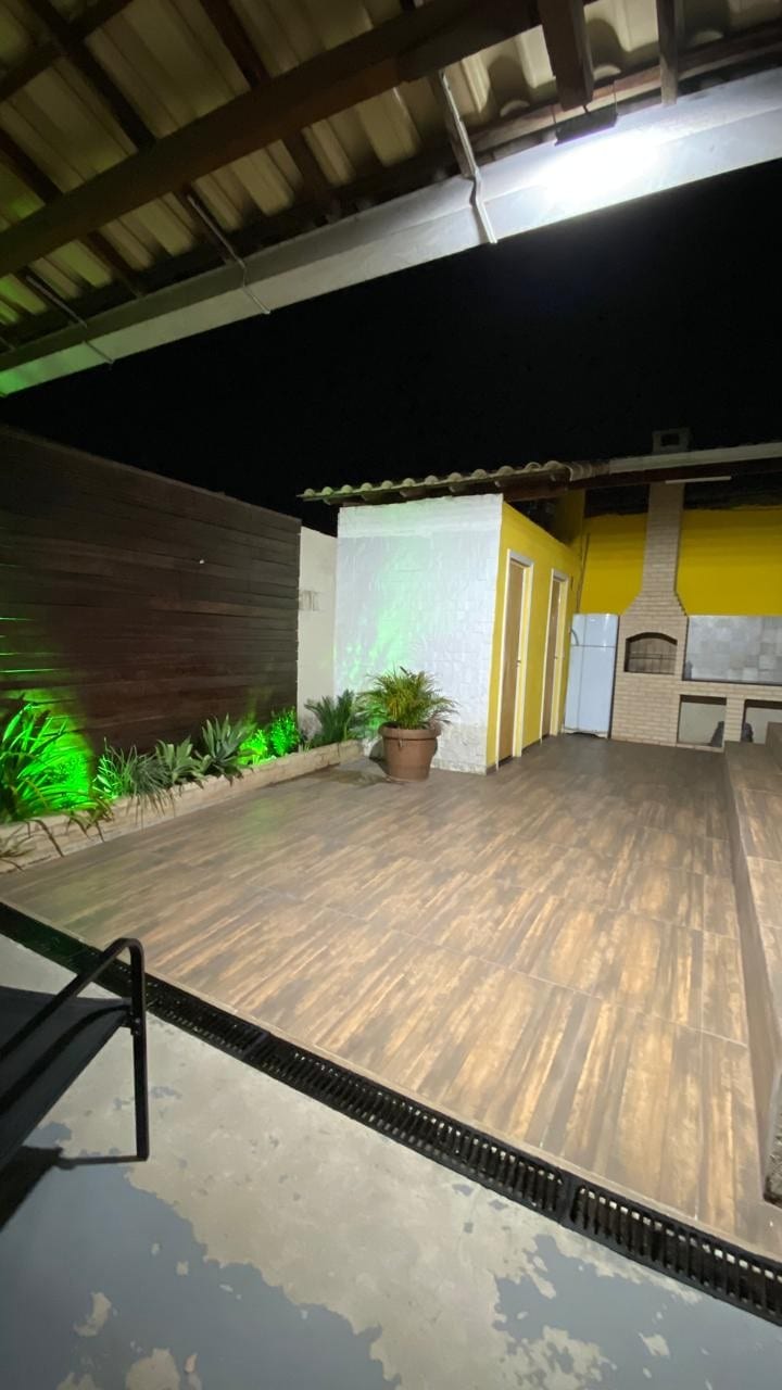 An outdoor area is showcased, featuring wooden flooring and green plants enhancing the space. The walls are painted in a bright yellow and white, with doors leading to separate rooms. A grill and kitchen area are visually present, positioned under a roof structure.