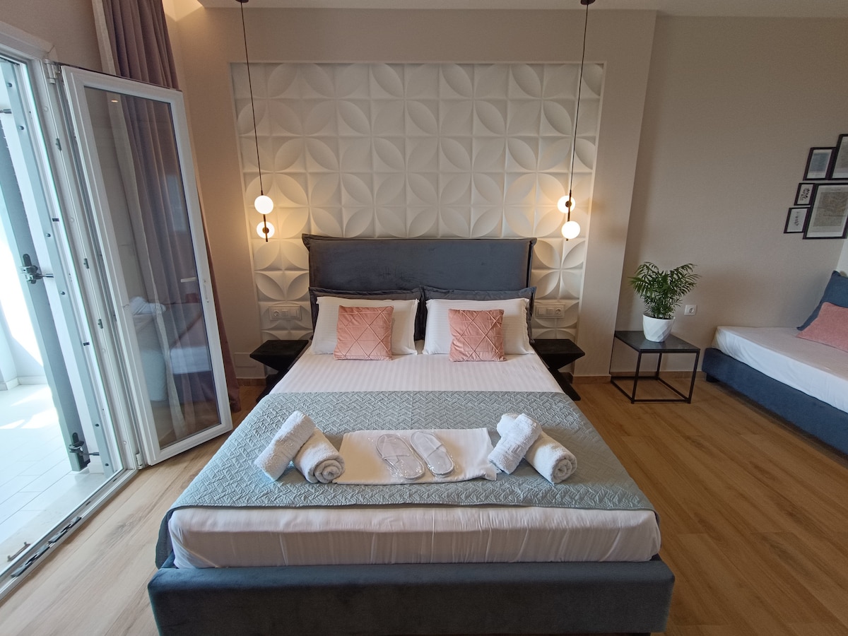 A spacious master bedroom is showcased, featuring a large bed with decorative pillows and neatly arranged towels on the sheets. Soft lighting is provided by two pendant lights. A balcony door opens to the outside, while a cozy sofa bed and a small table are visible in the background.