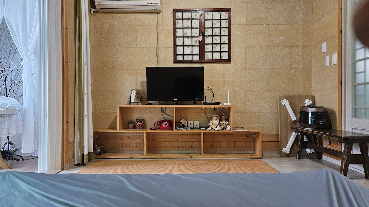 A living area features a mounted TV placed on a wooden console. The texture of the sandy walls is visible, complementing the simplicity of the space. A brown mat lies on the floor, creating a cozy atmosphere, while a small table and a heater are positioned nearby.