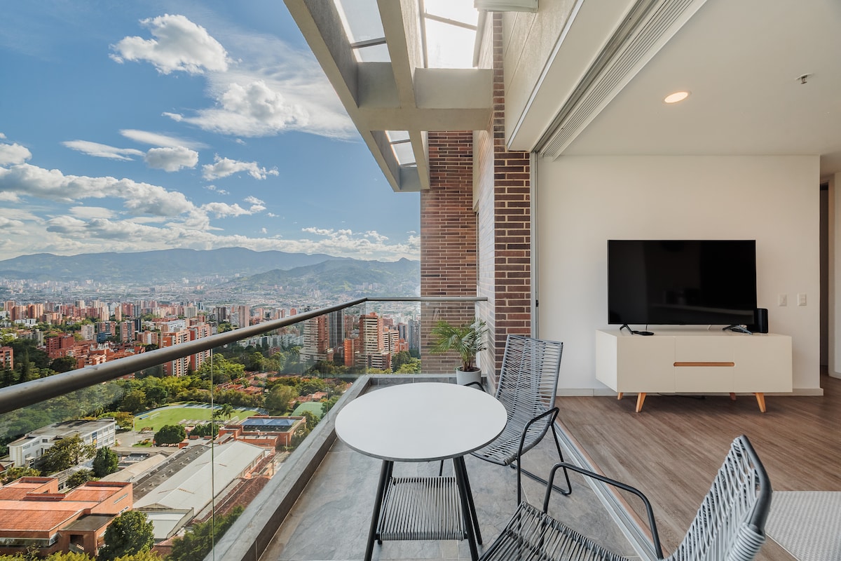 An outdoor balcony features a round table and two wire chairs, offering a comfortable space to enjoy the scenery. A large television sits on a minimalistic stand indoors, with expansive glass doors providing unobstructed views of the city and mountains in the background.