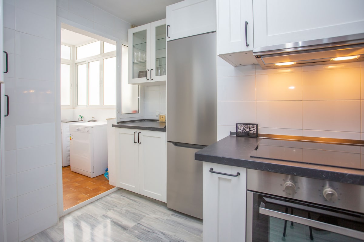 A modern kitchen is showcased, featuring sleek white cabinetry and stainless steel appliances. The countertop offers ample workspace, and a doorway leads to a bright laundry area with large windows. Natural light enhances the clean and functional design of the kitchen.