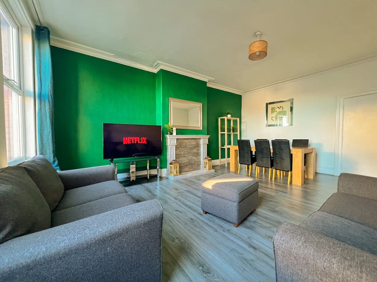 A bright living area features a green accent wall and large windows allowing natural light. Comfortable seating includes two gray sofas and an ottoman. A television is positioned in front of a decorative fireplace, with a dining table set for six nearby.