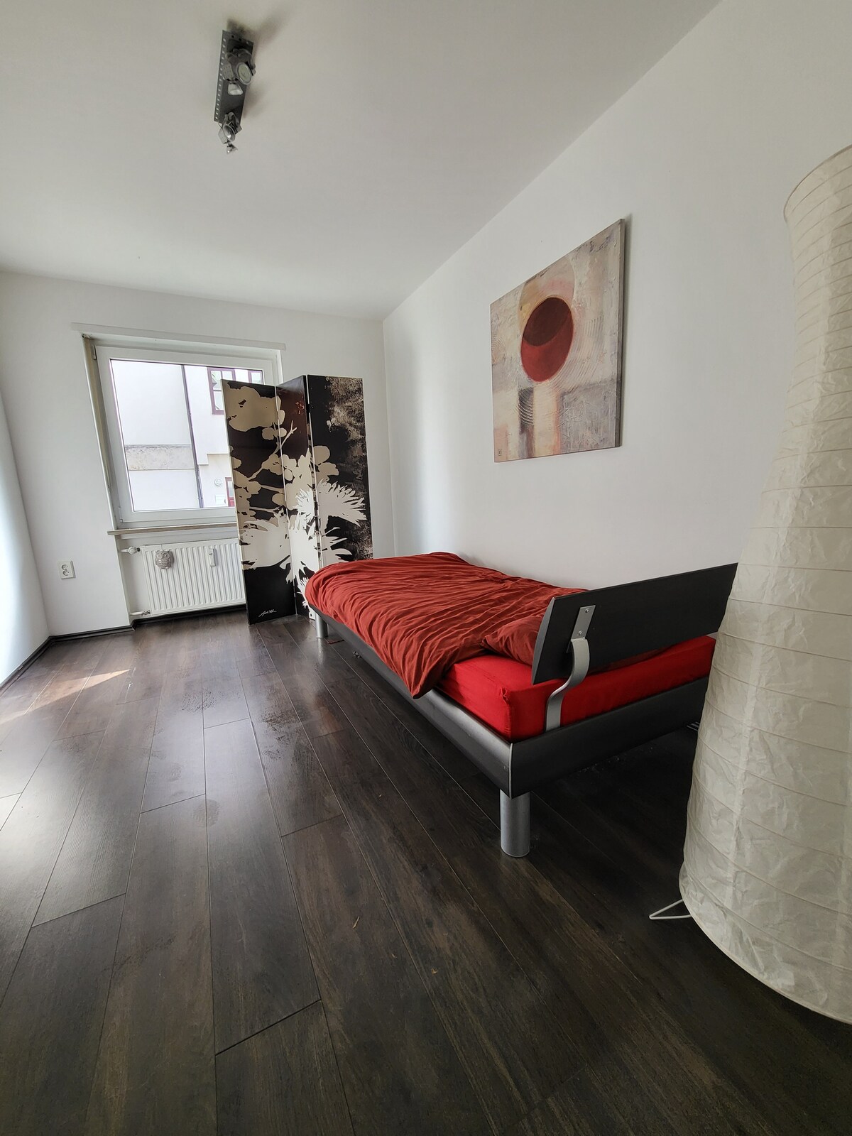 A cozy room features a single bed adorned with a red blanket. A decorative screen stands beside the bed, while a large window allows natural light to fill the space. Wooden flooring complements the contemporary design, and a floor lamp adds additional lighting.