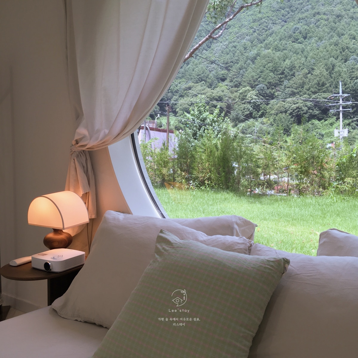 A cozy corner of the room is highlighted by a round window offering views of the surrounding forest. A soft lamp stands beside the bed, and a projector sits on the nightstand, suggesting options for entertainment. Gentle natural light filters through the soft curtains.