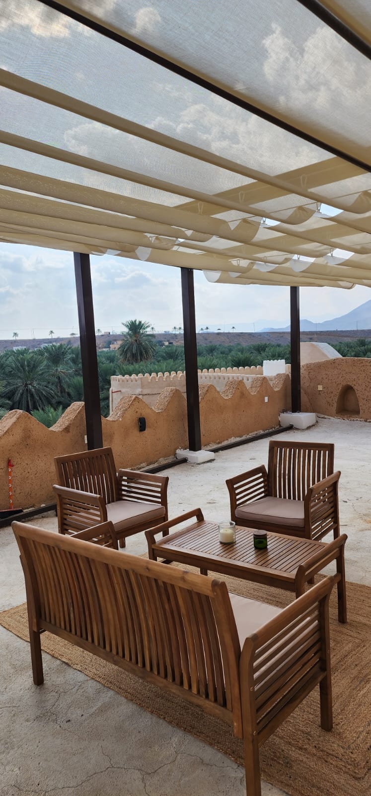 A serene rooftop seating area is featured, showcasing four wooden chairs with cushions surrounding a low table. The space is covered with a light fabric canopy, allowing natural light to filter through. Views of palm trees and mountains are visible in the background.