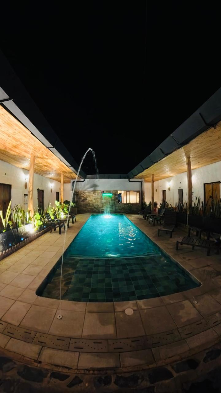 A serene swimming pool is bordered by well-maintained seating areas and illuminated by soft night lighting. A water feature creates a gentle arch over the pool, enhancing the tranquil atmosphere surrounded by greenery and colonial-style architecture.