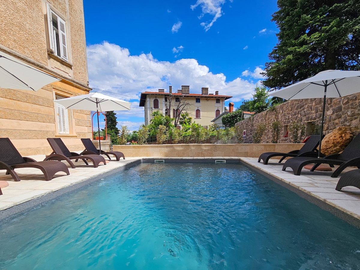 A tranquil pool area is framed by comfortable lounge chairs and umbrellas, providing shaded spots for relaxation. The water reflects the blue sky, while a stone wall and greenery enhance the serene environment. The villa's facade can be seen in the background.