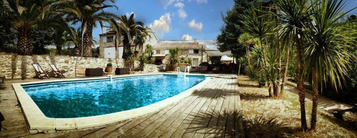 The image showcases a tranquil swimming pool surrounded by wooden decking and tropical plants. The clear blue water reflects the sky, enhancing the serene environment. Sun loungers are positioned nearby, offering a space for relaxation under the sun, with the rustic mas visible in the background.
