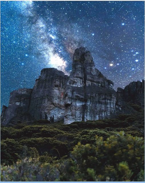 A stunning night sky filled with countless stars is displayed above a rugged stone mountain formation. The scene captures the towering cliffs illuminated softly by moonlight, surrounded by lush greenery at the base, showcasing the natural beauty of the landscape.