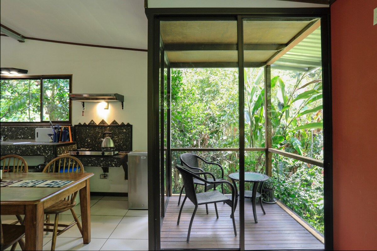 A view from inside the villa showcases a cozy dining space adjacent to a tastefully furnished outdoor balcony. Two chairs are positioned around a small table, surrounded by lush greenery visible through large glass doors, creating a seamless connection with nature.