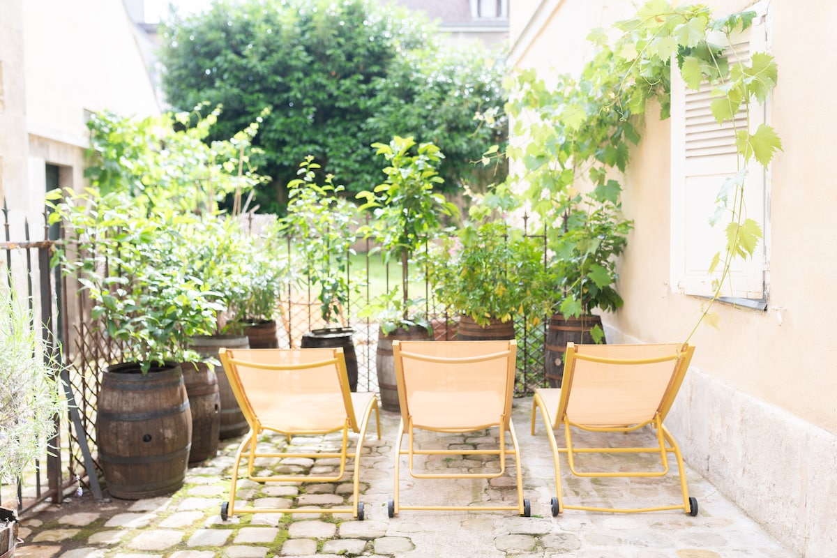 Three yellow lounge chairs are positioned on a stone patio, surrounded by lush greenery and potted plants. A low fence made of decorative ironwork encloses the area, and sunlight casts a warm glow on the vibrant foliage.