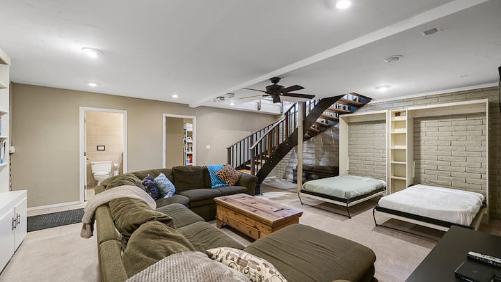 מרתף רב-תכליתי עם ספה גדולה, קיר לבנים, 2 מיטות נפתחות וגרם מדרגות מעוקל.