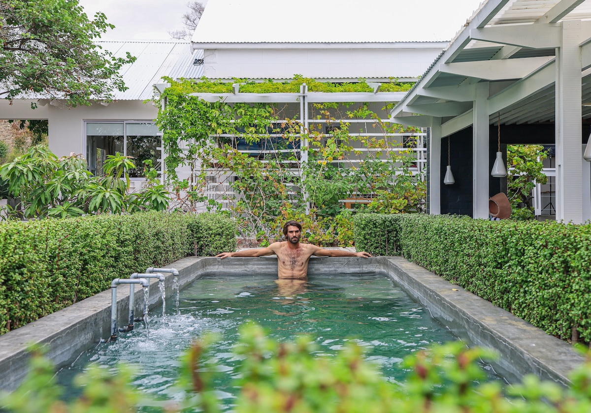 A serene swimming pool is surrounded by neatly trimmed greenery, providing a tranquil atmosphere. The water reflects the surrounding landscape, while a person is seen relaxing at the edge of the pool, creating a sense of peace and privacy.