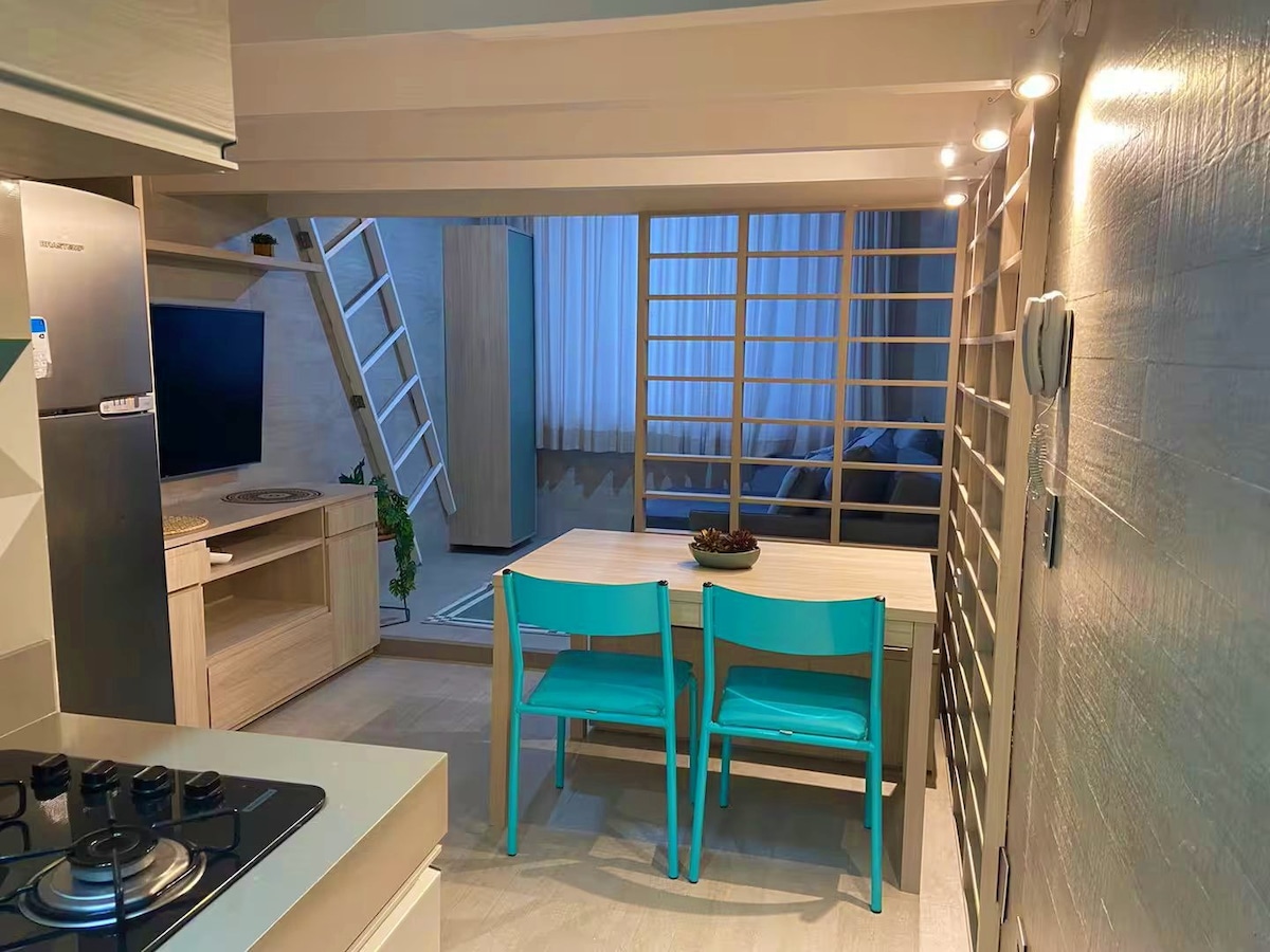 A dining area is visible with a light wooden table and two turquoise chairs, creating a bright and modern atmosphere. The kitchen area is equipped with a stove and refrigerator, while a ladder leads to the sleeping loft above, enhancing the vertical space.