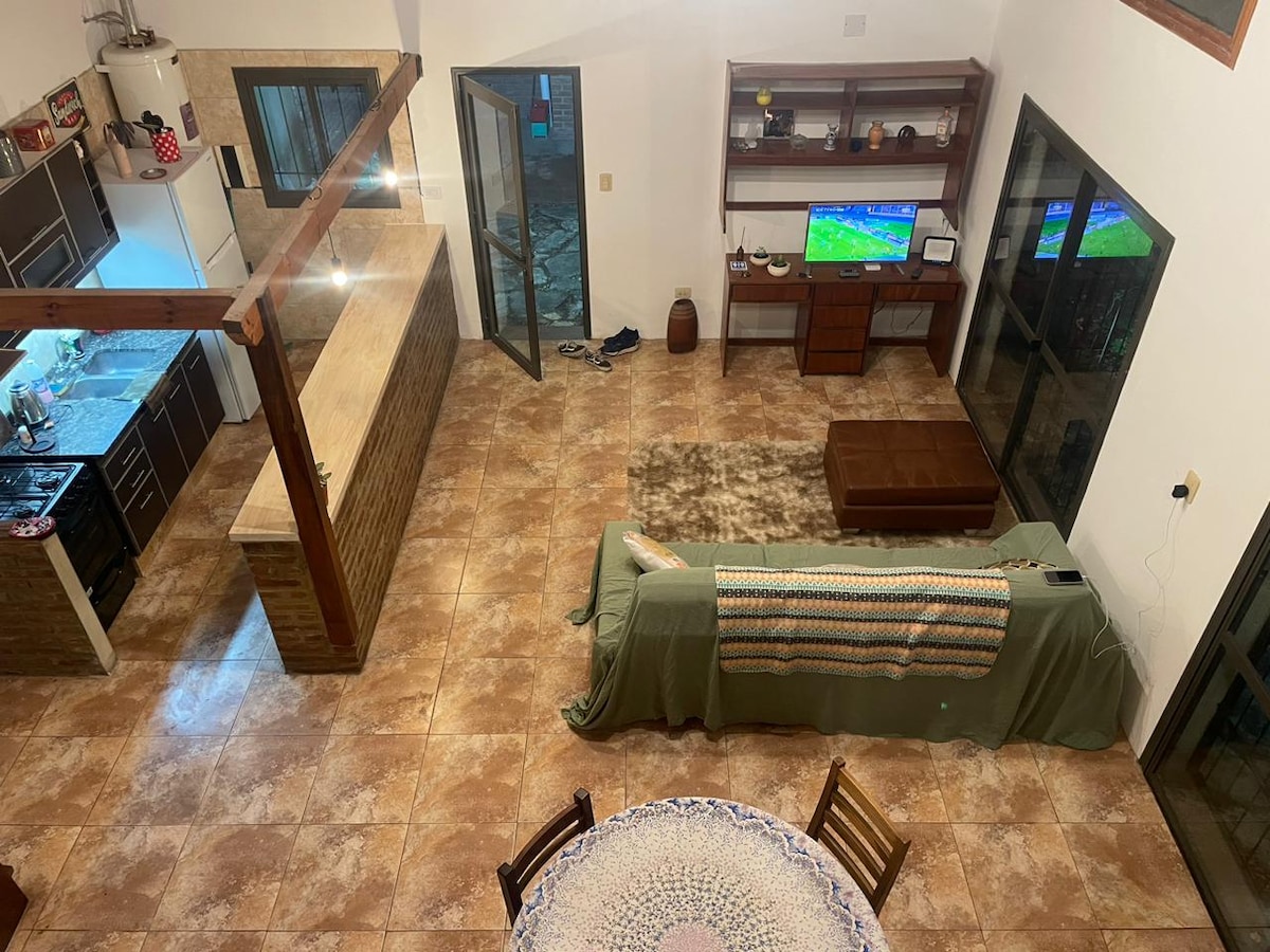An open-concept living area with a view of a spacious kitchen and dining area. A brown coffee table is placed in front of the sofa, which is draped with a green blanket. Large windows allow natural light to fill the room, enhancing the inviting atmosphere.