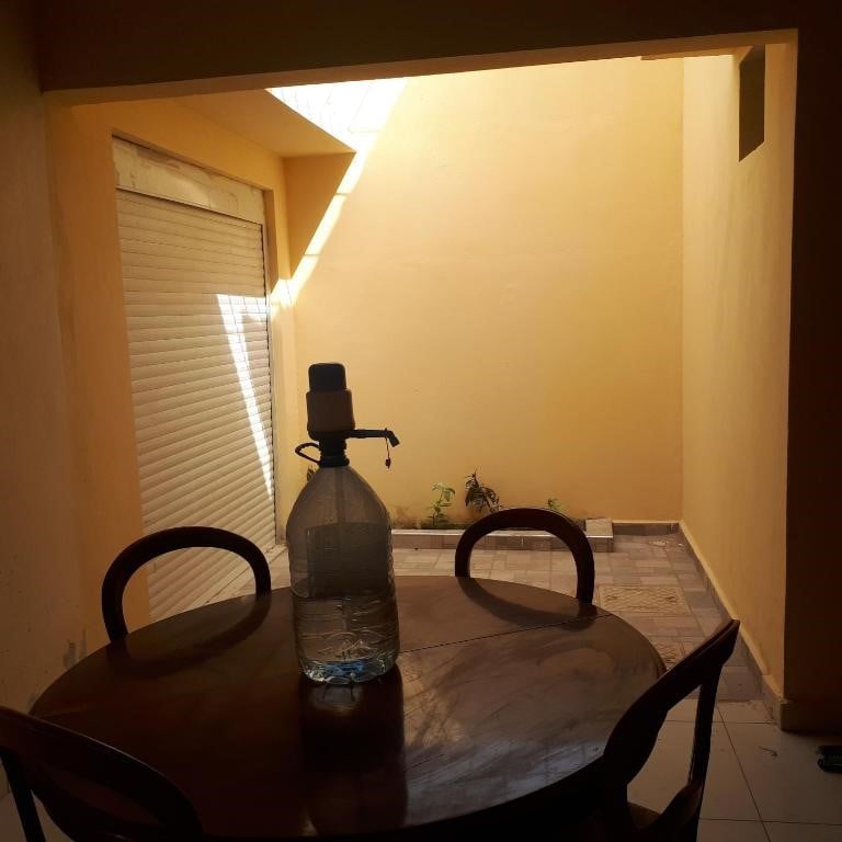 An indoor space features a small wooden table with two chairs. A large glass bottle is placed on the table. Natural light is observed streaming in from an open area, illuminating the yellow-walled surroundings and a glimpse of green plants in the background.