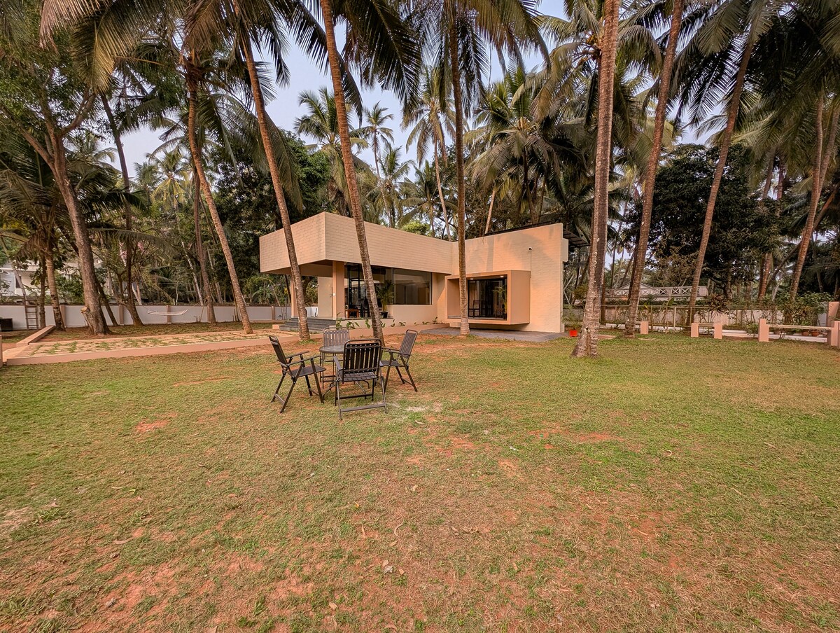 The cottage is set within a spacious outdoor area, surrounded by palm trees. An inviting lawn features a circular arrangement of outdoor furniture, providing a comfortable space for relaxation and gatherings. Large glass windows reflect the natural surroundings, enhancing the serene setting.