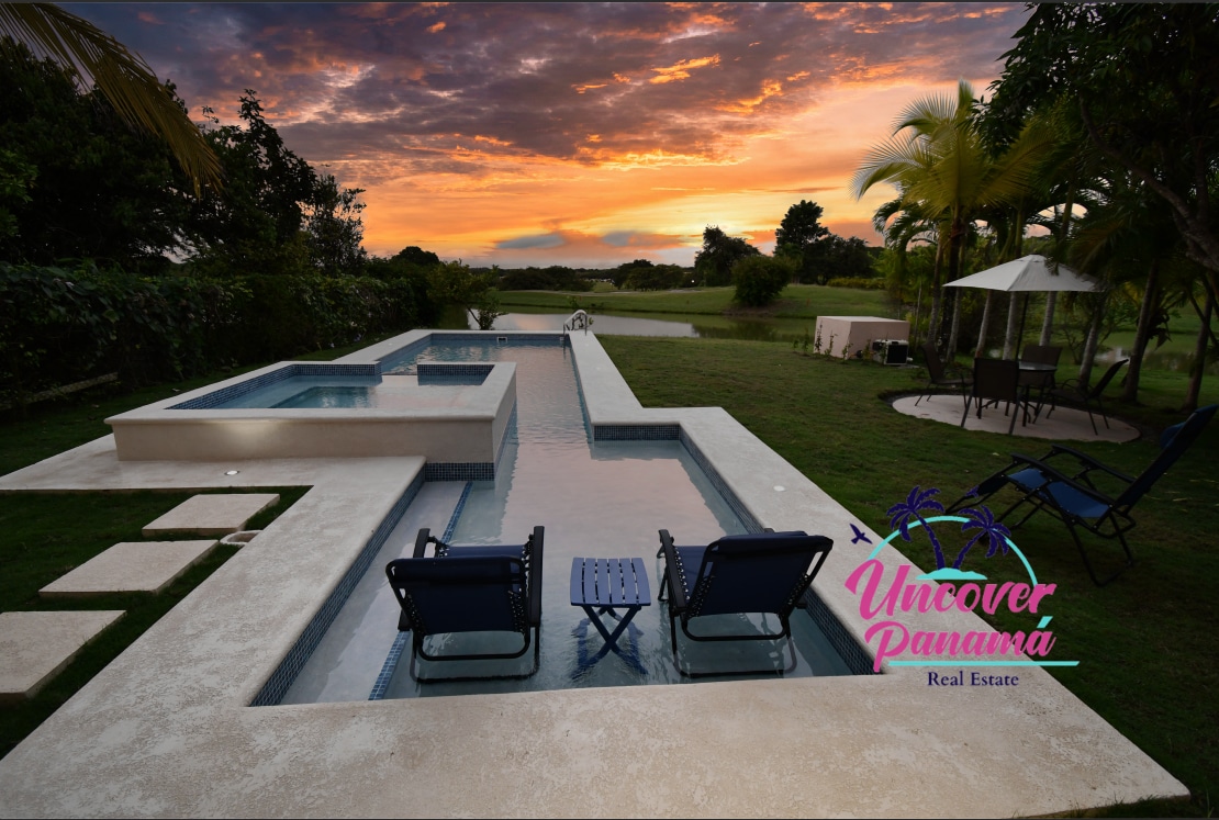 A serene outdoor space features a long swimming pool alongside a manicured lawn. Two loungers are positioned at one end of the pool, offering a view of the sunset and surrounding trees. The sky showcases hues of orange and pink as the day wanes.