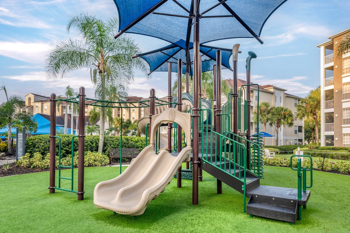 A children's playground is displayed with colorful slides and climbing structures shaded by large blue canopies. Lush green grass surrounds the play area, while palm trees and neighboring buildings are visible in the background.