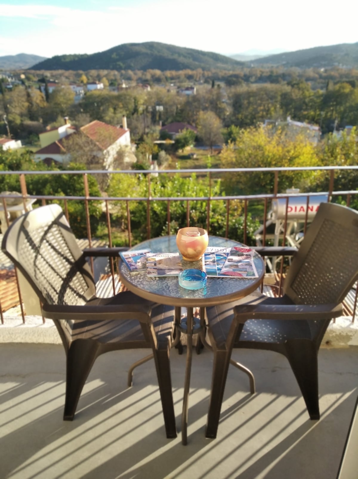 A small balcony table is set with a drink and magazines, flanked by two chairs. In the background, a panoramic view of the village and surrounding hills can be seen, bathed in natural light.