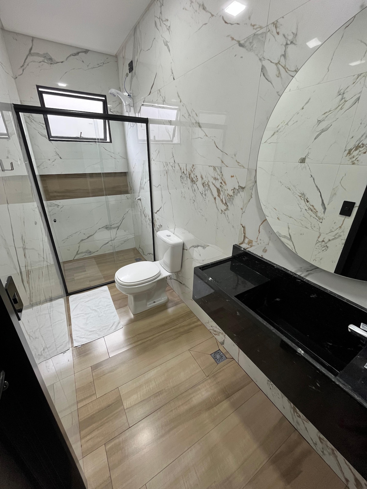 A modern bathroom features elegant marble walls, a spacious shower area with glass doors, and a sleek black countertop with a large mirror. Lighting fixtures provide ample illumination, while wooden floor tiles create a warm atmosphere. Essential bathroom fixtures are present, ensuring functionality.