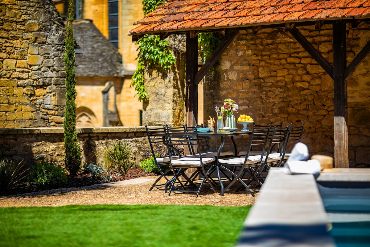 An outdoor dining area features a large table set with fresh flowers and fruit. Black metal chairs surround the table in a shaded spot beneath a wooden pergola. Stone walls and lush greenery frame the serene space, with a pool visible in the foreground.