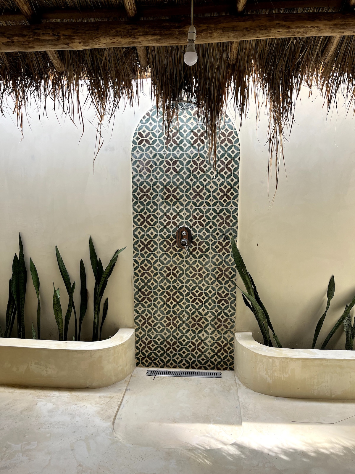 An outdoor shower is framed by a decorative tiled arch, highlighted by earthy tones. Tall, leafy plants are positioned along the sides of the shower, enhancing the natural setting. A thatched roof overhead provides shade while a single light bulb adds a touch of functionality.
