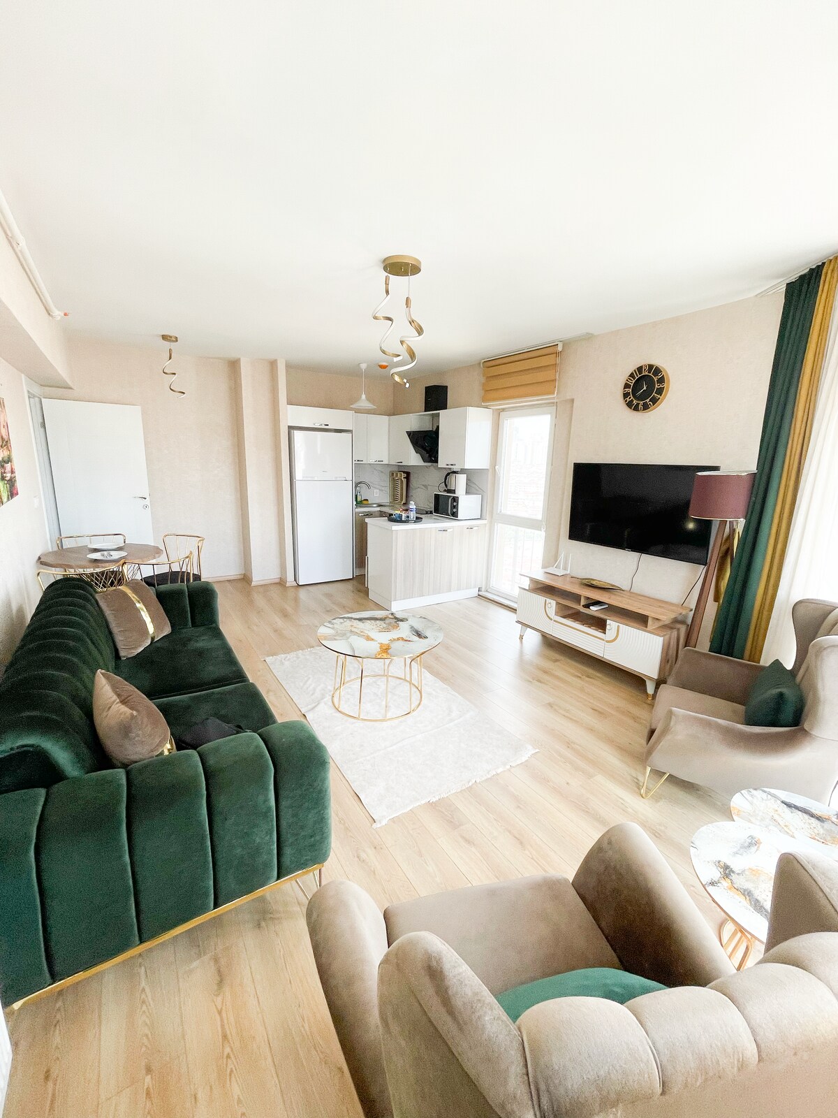 A well-furnished living area with a green velvet sofa and two accent chairs offers a comfortable seating arrangement. A small glass coffee table is positioned in front, while a kitchenette is visible in the background, equipped with modern appliances. Natural light floods the space through large windows.